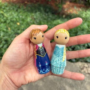 May include: Two wooden peg dolls, one with brown hair and a blue and pink dress, the other with blonde hair and a blue dress. The dolls are held in a hand against a blurred green background.