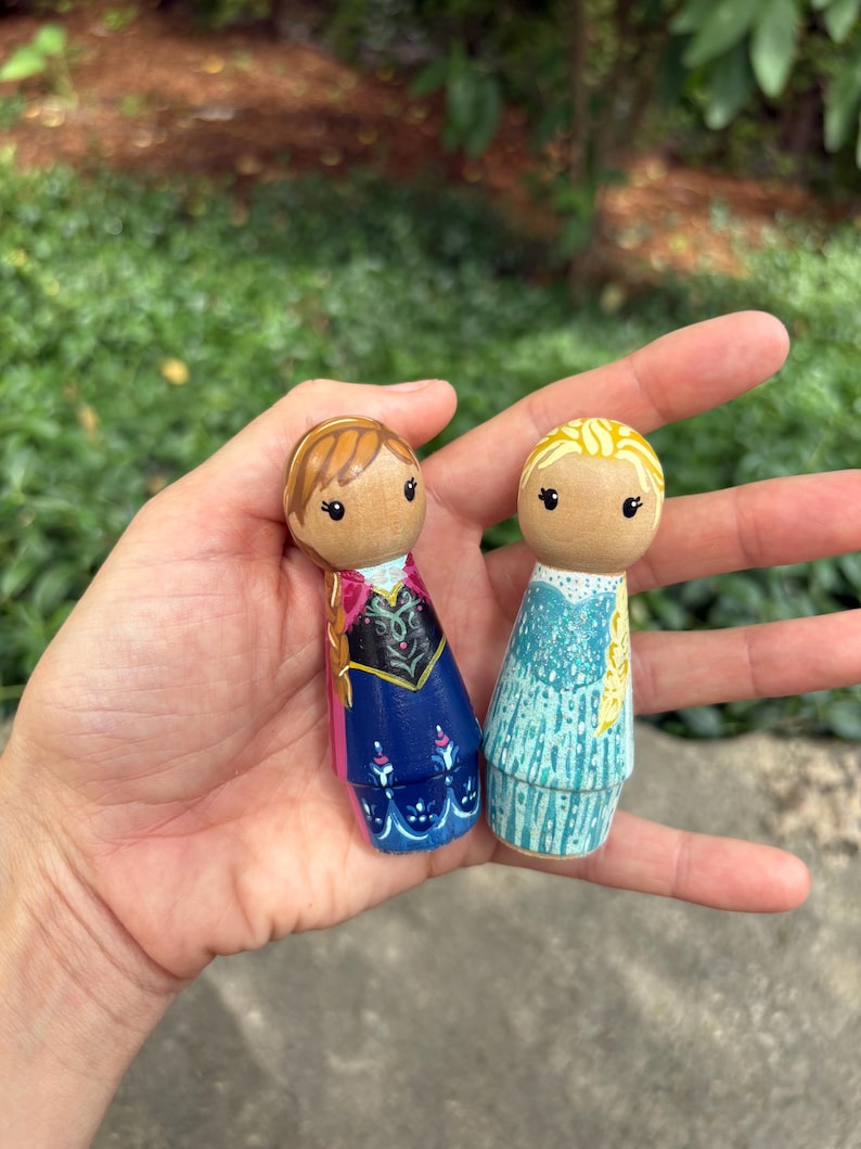 May include: Two wooden peg dolls, one with brown hair and a blue and pink dress, the other with blonde hair and a blue dress. The dolls are held in a hand against a blurred green background.