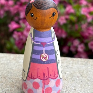 May include: A hand-painted wooden peg doll depicting a character in a white lab coat, purple striped shirt, and pink skirt with polka dots. The doll has brown skin, dark hair, and a pink flower. A red heart-shaped pendant hangs from the doll's neck.