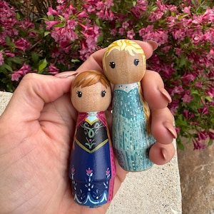 May include: Two hand-painted wooden peg dolls, one with brown hair and a blue and black dress, the other with blonde hair and a blue dress. The dolls have detailed facial features and clothing designs. The background features vibrant pink flowers.
