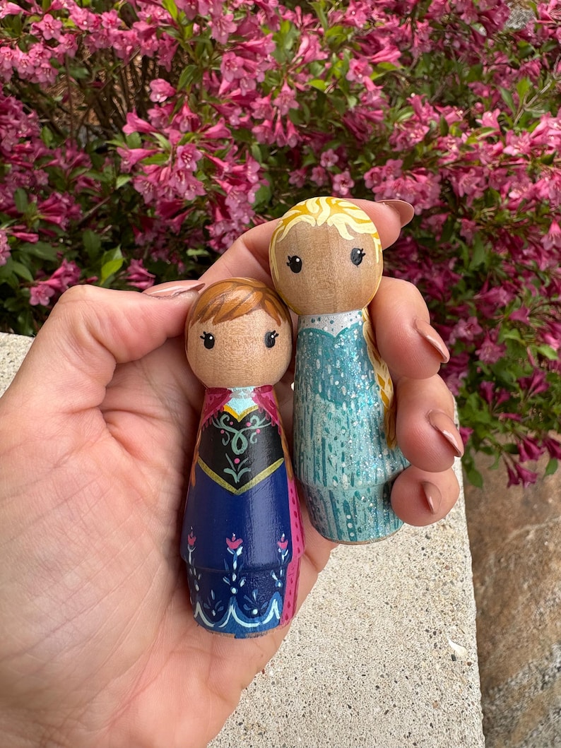 May include: Two hand-painted wooden peg dolls, one with brown hair and a blue and black dress, the other with blonde hair and a blue dress. The dolls have detailed facial features and clothing designs. The background features vibrant pink flowers.