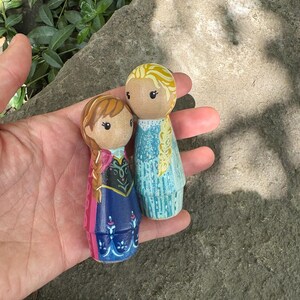 May include: Two hand-painted wooden peg dolls, depicting characters from a popular animated film. One doll has blonde hair in a braid and a blue dress, the other has brown hair and a blue and pink dress.