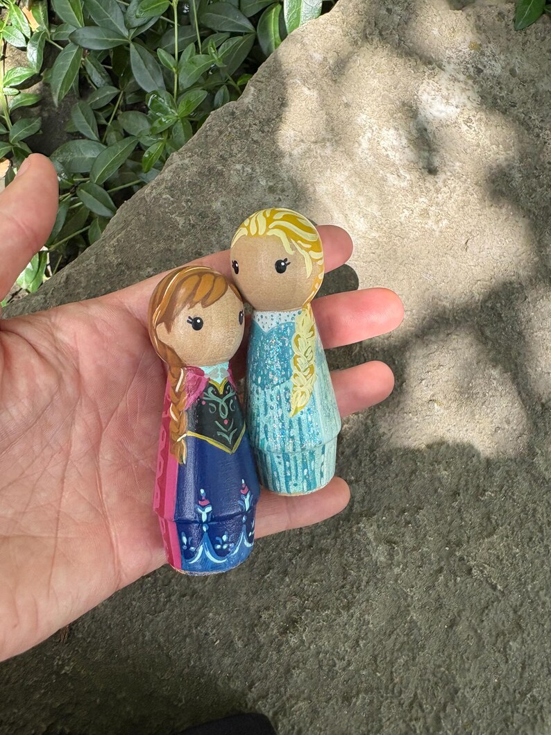 May include: Two hand-painted wooden peg dolls, depicting characters from a popular animated film. One doll has blonde hair in a braid and a blue dress, the other has brown hair and a blue and pink dress.