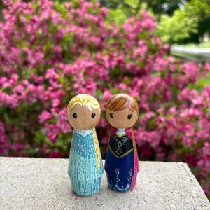 May include: Two wooden peg dolls, one in a light blue dress with blonde hair and the other in a dark blue dress with brown hair. The dolls are standing on a concrete surface with a backdrop of pink flowers.