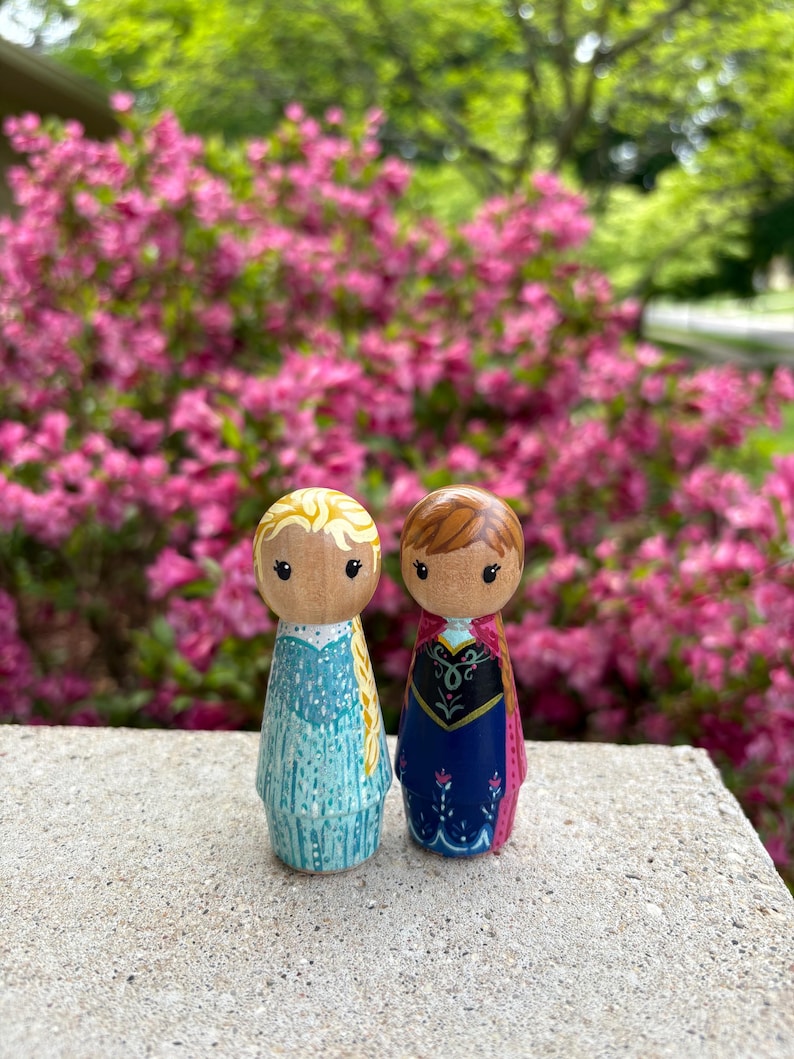 May include: Two wooden peg dolls, one in a light blue dress with blonde hair and the other in a dark blue dress with brown hair. The dolls are standing on a concrete surface with a backdrop of pink flowers.