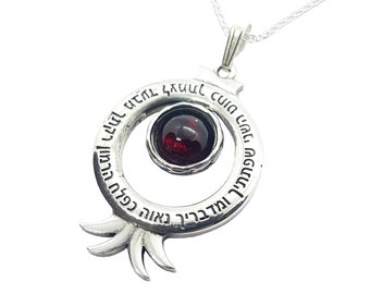 May include: Silver necklace with a pomegranate pendant featuring a red gemstone and Hebrew text. The text reads "May you be blessed with many descendants like the seeds of a pomegranate."