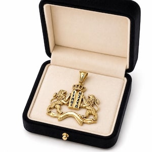 May include: A gold-colored pendant featuring two lions flanking a tablet with Hebrew text, set within a black velvet jewelry box. The pendant has a crown design above the tablet and a decorative banner below the lions.
