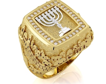 May include: A gold-colored ring with a square face featuring a silver menorah design set against a textured gold background. The face is bordered by small, clear gemstones. The ring's band is detailed with a city scene.