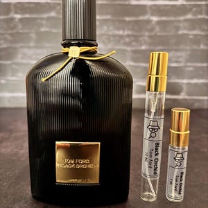 Op de afbeelding: Een zwarte glazen fles Tom Ford Black Orchid parfum met een gouden label en een zwarte geribbelde dop. Twee kleinere flesjes van hetzelfde parfum zijn ook te zien, een van 10 ml en de andere van 5 ml.