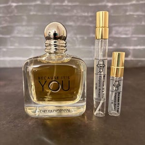 Pode incluir: Um frasco de perfume de vidro transparente com uma tampa dourada. O frasco é etiquetado com "Because It's You" e "Emporio Armani". Há duas garrafas de perfume menores ao lado da garrafa maior.