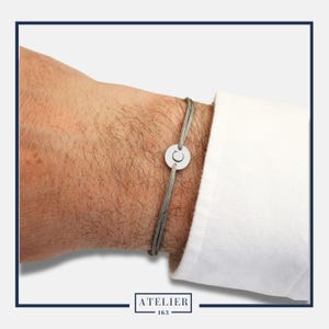 Peut inclure: Un bracelet argenté avec un charm circulaire représentant un croissant de lune. Le bracelet est composé d'un cordon gris et est porté au poignet. L'image montre également une manche de chemise blanche. La marque "ATELIER 163" est en bas.