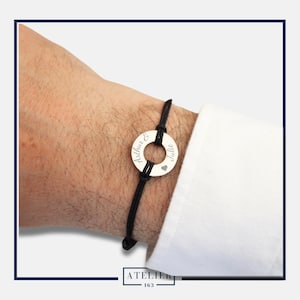 Peut inclure: Un bracelet en cordon noir avec un charm circulaire argenté. Le charm porte les noms "Arthur & Julie" gravés, ainsi qu'un petit symbole de cœur. Le bracelet est porté sur un poignet, avec un poignet de chemise blanche visible.