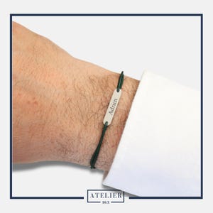 Peut inclure: Un bracelet en cordon vert avec une plaque argentée portant le nom "Adrien". Le bracelet est porté sur un poignet, avec un revers de chemise blanche visible. Le texte "ATELIER 163" est affiché en bas de l'image.