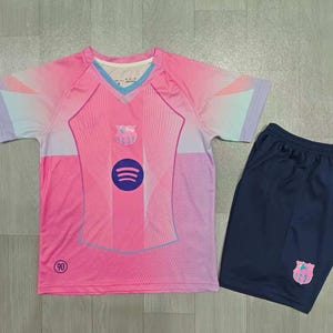 Può includere: Una maglia a maniche corte rosa e azzurro con scollo a V, caratterizzata da un logo Spotify centrale, uno stemma sul petto e il numero 90. Abbinata a pantaloncini blu scuro con uno stemma corrispondente sulla parte inferiore della gamba.