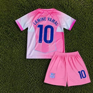 Peut inclure: Un ensemble de football rose et blanc composé d'un maillot et d'un short. Le maillot porte le nom LAMINE YAMAL et le numéro 10 en bleu au dos. Le short présente le numéro 10 en bleu et un petit écusson d'équipe sur le tissu rose.