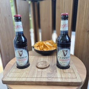 Könnte beinhalten: Zwei Flaschen Guinness Extra Stout mit roten Kronkorken stehen auf einem Holzservierbrett. Ein kleiner Holzuntersetzer und eine Schüssel mit Chips befinden sich ebenfalls auf dem Tisch. Die Flaschen haben weiße Etiketten mit dem Guinness-Logo.