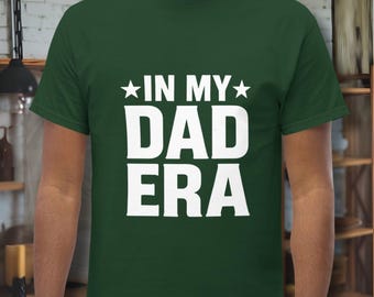 Peut inclure: Un t-shirt vert foncé avec le texte blanc "IN MY DAD ERA". Le texte est en gras, avec les mots "IN MY" au-dessus de "DAD ERA". Le t-shirt a des manches courtes et un col rond.