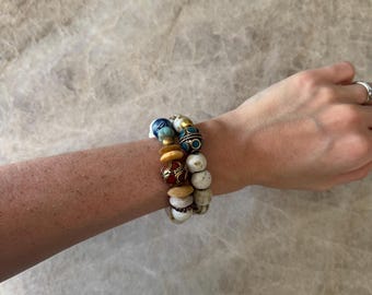 Op de afbeelding: Een armband met meerdere strengen en diverse kralen in verschillende vormen, maten en kleuren, waaronder wit, beige, blauw en een donkerrode kraal met goudkleurige metalen accenten, gedragen om de pols met een eenvoudige zilverkleurige ring.