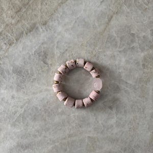 Puede incluir: Una pulsera de cuentas rosas con una sola cuenta de cuarzo rosa. La pulsera presenta cuentas cilíndricas con una textura moteada y espaciadores de color latón. La cuenta de cuarzo rosa tiene forma ovalada lisa.
