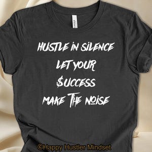 May include: A dark grey t-shirt with white text that reads "Hustle in Silence Let Your Success Make the Noise". The text is in a bold, sans-serif font. The shirt has a crew neck and short sleeves. The bottom of the shirt has the text "Happy Hustler Mindset" in a smaller font.