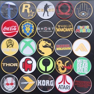 May include: A collection of 25 round embroidered patches featuring various video game, movie, and pop culture logos. The patches are arranged in a grid pattern on a black background. Some of the logos include Star Trek, Coca-Cola, Ghost of Tsushima, World of Warcraft, Super Mario, Thor, Metal Gear Solid, and Stranger Things.