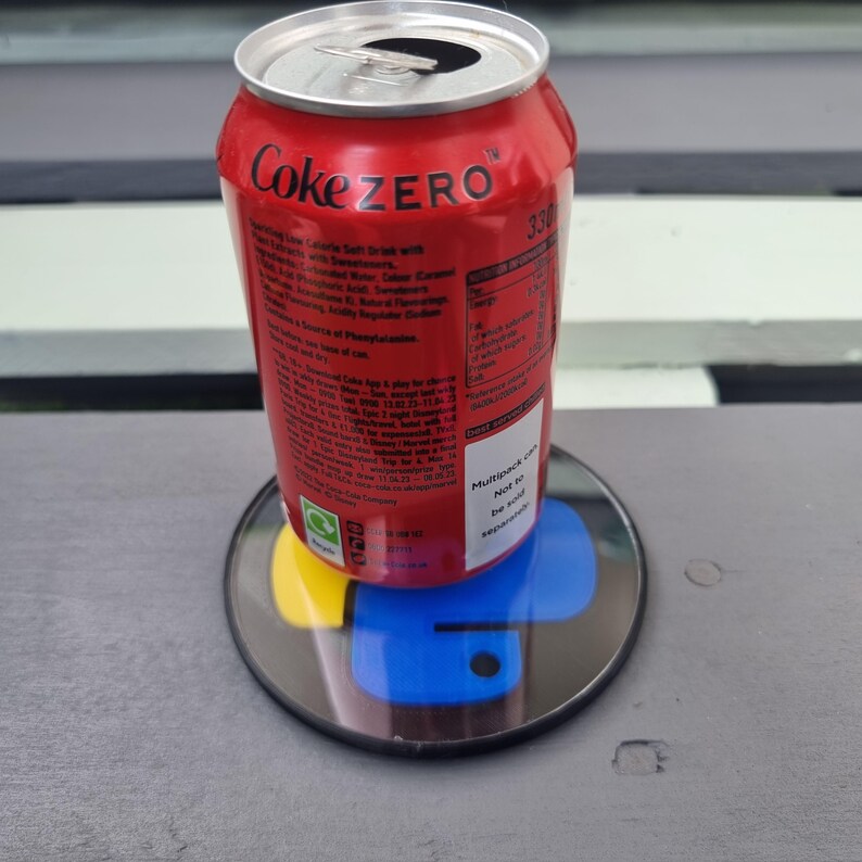 May include: A red aluminum can of Coke Zero with white lettering. The can is sitting on a colorful round coaster with a black border. The can has nutritional information printed on the side.