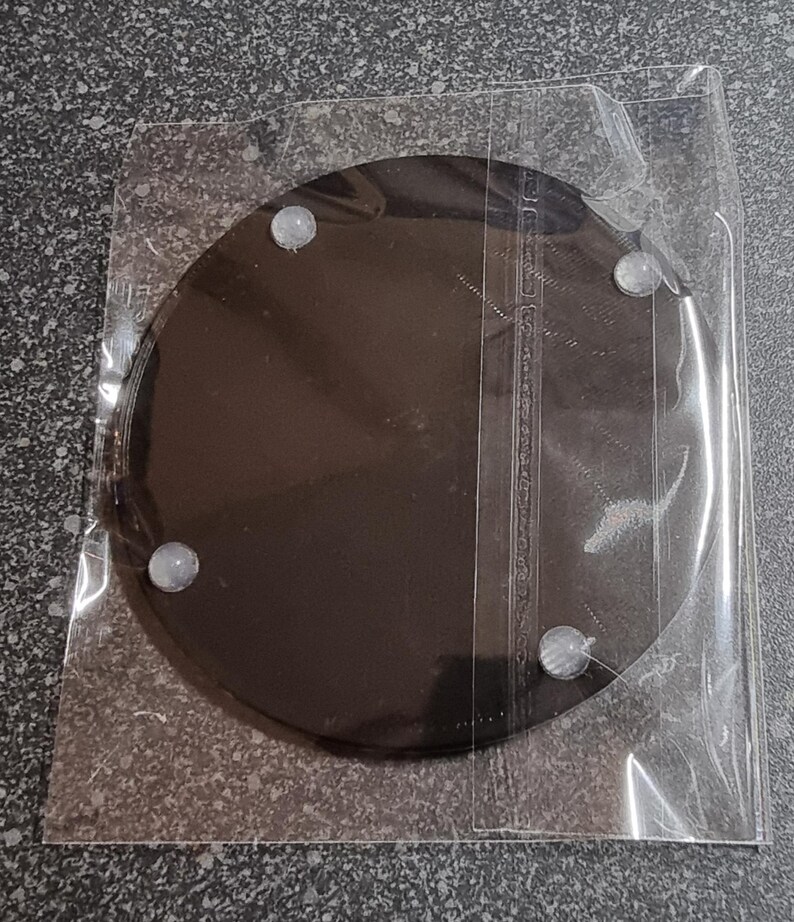 May include: A black round coaster with four white rubber feet on the bottom. The coaster is packaged in a clear plastic bag.