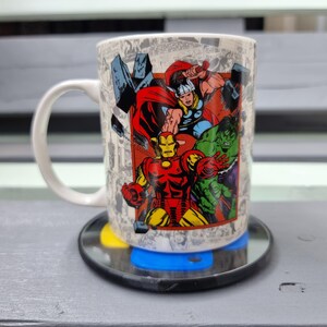 May include: White ceramic mug with a comic book style image of Iron Man, Thor, and the Hulk. The mug has a black handle and is sitting on a black coaster.