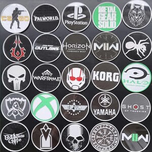 May include: A collection of 20 round, black and white stickers with various video game, movie, and brand logos. The logos include: CS:GO, Palworld, PlayStation, Metal Gear Solid, Horizon Forbidden West, Korg, Halo, Warframe, Ghost of Tsushima, Yamaha, Top Gun, MIN, Outlaws,  and more.