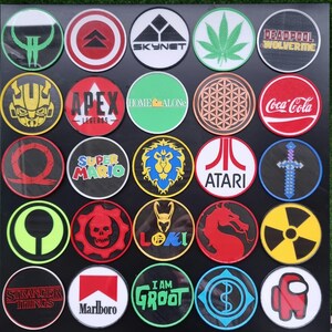 May include: A collection of 25 round iron-on patches featuring various logos and designs. The patches are colorful and include logos such as Coca-Cola, Apex Legends, Super Mario, and Stranger Things.