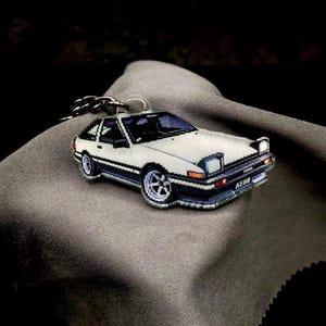 Op de afbeelding: Een wit en zwarte autosleutelhanger met blauwe accenten. De sleutelhanger heeft een gedetailleerd ontwerp van een klassiek automodel, compleet met wielen en koplampen. De sleutelhanger is bevestigd aan een zilveren ketting.