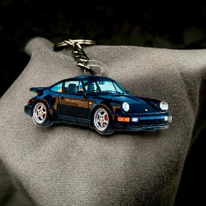 Op de afbeelding: Een zwarte autosleutelhanger met zilveren accenten en een sleutelring. De sleutelhanger is een miniatuurreplica van een sportwagen, met gedetailleerde wielen en ramen. De auto staat tegen een grijze stoffen achtergrond.