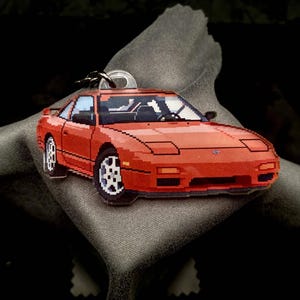 Op de afbeelding: Een gepixelde rode sportwagen sleutelhanger met witte wielen en een zilveren sleutelring. De auto heeft zwarte ramen en koplampen. De sleutelhanger ligt op een grijze, getextureerde ondergrond tegen een zwarte achtergrond.
