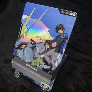 May include: A holographic trading card featuring anime characters and a robot. The card has the text "EX Base" and "EXB-028". The background is blue with a futuristic design. The card is displayed on a clear stand.
