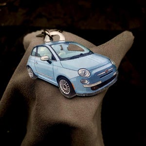Op de afbeelding: Een lichtblauwe autosleutelhanger met een zilveren sleutelring. De sleutelhanger is een miniatuurreplica van een klassieke auto, met gedetailleerde ramen, wielen en een grille. De auto ligt op een grijze ondergrond tegen een donkere achtergrond.