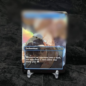 Może przedstawiać: Prostokątna karta kolekcjonerska z ilustracją postaci i tekstem Enchantment. Whenever an opponent casts a spell, you may draw a card unless that player pays 1. Karta jest umieszczona na małej, przezroczystej plastikowej podstawce.