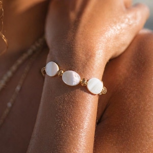 May include: A gold bracelet featuring three oval, white mother-of-pearl stones. The bracelet is worn on a wrist, with a gold necklace visible. The image is a close-up, with soft focus and natural lighting.