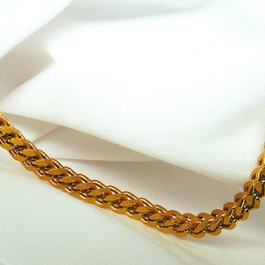 May include: A gold-colored chain necklace with a woven design. The chain is displayed against a white fabric background, creating a contrast that highlights the necklace's texture and color.