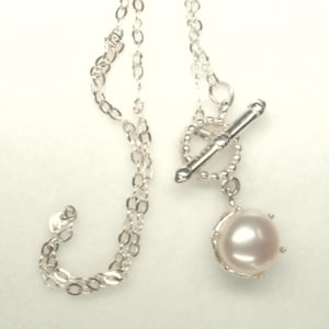May include: A silver necklace with a toggle clasp and a single, round, light pink pearl pendant. The chain is made of small, linked silver pieces. The pearl is set in a decorative silver setting.