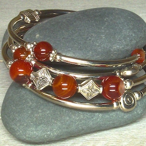 May include: A set of silver-toned bangles with reddish-brown beads and decorative silver accents. The beads are interspersed with silver floral and geometric designs. The bangles are stacked on a gray stone.