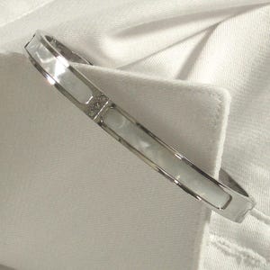 May include: A silver-toned bangle bracelet with rectangular mother-of-pearl inlays and small, sparkling accents. The bracelet is shown against a white background, highlighting its elegant design and reflective surface.