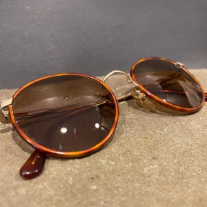 May include: Vintage round sunglasses with a tortoiseshell pattern on the frames. The lenses are a dark brown color, and the arms are a dark brown. The sunglasses are resting on a stone surface.
