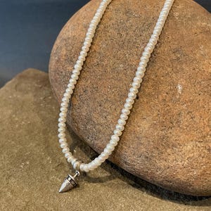 May include: A white pearl necklace with a silver pendant shaped like a pointed cone. The necklace is on a brown rock.