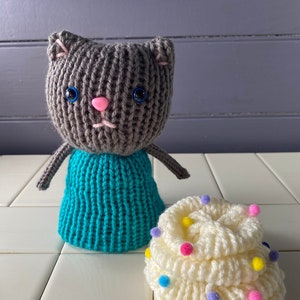 May include: A gray knitted cat doll wearing a teal skirt stands next to a white knitted cupcake with colorful sprinkles.