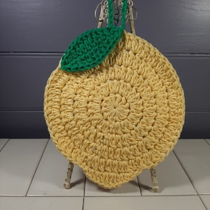 May include: A crocheted lemon-shaped pot holder with a green leaf. The pot holder is yellow and is displayed on a decorative stand.