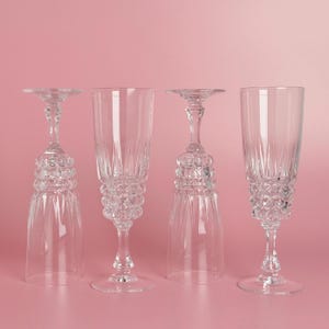 May include: Four clear champagne flutes with detailed stem and base designs are set against a pink backdrop. Two flutes are upright, and two are inverted, highlighting the glassware's intricate craftsmanship.