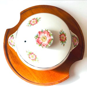 May include: White ceramic casserole dish with a floral design featuring pink and white flowers. The dish has a lid with a knob in the center and two handles on the sides. The dish is sitting on a wooden base.