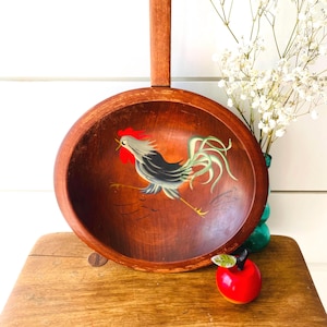 May include: A round, wooden bowl with a handle, featuring a hand-painted rooster design in the center. The rooster has red, gray, and green accents. The bowl is brown and sits on a wooden surface.