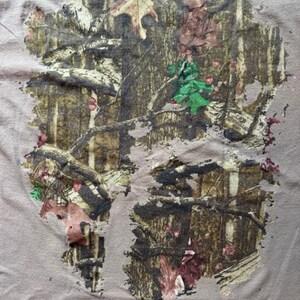 Op de afbeelding: Een bruin T-shirt met een camouflagepatroon bedrukt op de voorkant. Het patroon is in de kleuren bruin, groen en wit.