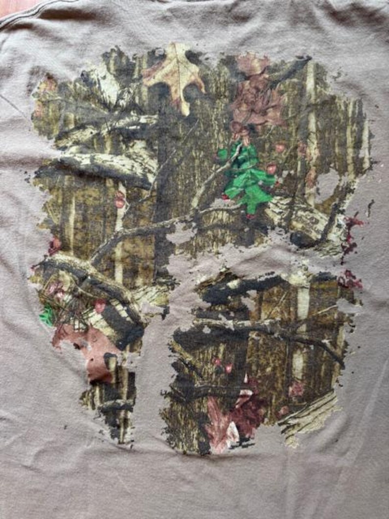 Op de afbeelding: Een bruin T-shirt met een camouflagepatroon bedrukt op de voorkant. Het patroon is in de kleuren bruin, groen en wit.